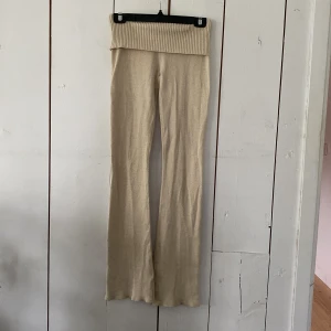 Beiga stickade byxor från Gina Tricot - Mysiga beiga stickade byxor från Gina Tricot med bred ribbad midja som kan vikas ner. Byxorna har en rak och lite utsvängd passform, perfekta för en chill och avslappnad stil. Supermjuka och sköna att ha på sig hela dagen.