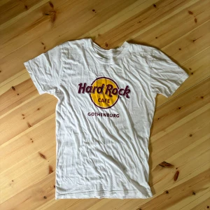 Hard Rock Cafe t-shirt Göteborg - Vit t-shirt från Hard Rock Cafe med klassiskt tryck i gult och vinrött på bröstet och texten 'Gothenburg' under loggan. T-shirten har rund hals och korta ärmar, perfekt för dig som gillar ikoniska bandtröjor och streetstyle. Lite skrynklig men är bara tvätta den eller stryka den.