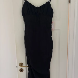 Svart långklänning från Nelly - Säljer en svart långklänning från Nelly, modell Catch My Eye Dress. Klänningen har smala axelband, snygg snörning framtill och ett krinklat tyg som ger cool struktur. Passformen är slim och klänningen har en elegant siluett som framhäver figuren.