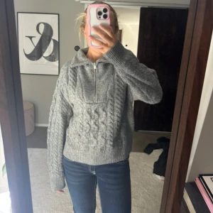 Grå stickad tröja med half zip - Säljer en grå stickad tröja med snygg kabelstickning och half zip framtill. Tröjan har hög krage och långa ärmar, perfekt för kyliga dagar. Ribbstickade muddar vid ärmslut och nederkant.🥰