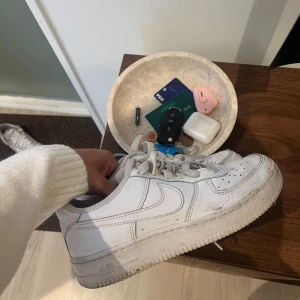 Vita Nike Air Force 1 sneakers - Klassiska vita Nike Air Force 1 sneakers med låg profil och rund tå. Skorna har snörning och är tillverkade i skinn med en platt sula. Perfekta för dig som gillar streetstyle och vill ha en ikonisk sneaker i garderoben.