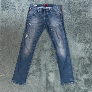Blå slitna jeans från Jack & Jones - Säljer ett par blå jeans från Jack & Jones med slitningar och lätt tvättad look. Storlek 33/32. Byxorna har fem fickor, dragkedja och knapp i midjan. Skinny/slim passform och normal midja. Perfekta för en avslappnad stil.