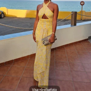 Gul långklänning med omlott och mönster - Säljer en superfin gul långklänning med ljusgrått blommigt mönster. Klänningen har omlott-design upptill med snygg cut-out vid midjan och korsade axelband. Materialet är lätt och luftigt, perfekt för varma dagar. Passformen är slim och klänningen är ärmlös.