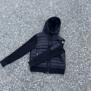 Svart Moncler cardigan  - Säljer en svart pufferjacka från Moncler med stickade ärmar och ribbade muddar. Jackan har quiltad kropp, dragkedja framtill och en stor huva. Perfekt för dig som vill ha en snygg och varm jacka med streetkänsla.