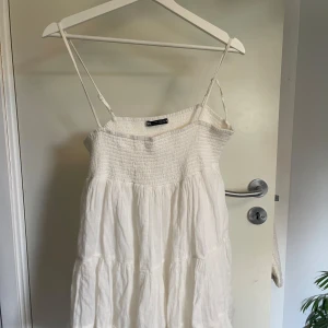 Vit sommarklänning med smala axelband - Supersöt vit klänning från Zara med smala axelband och smock upptill. Klänningen har en luftig och flowy passform med volanger och är perfekt för varma dagar. Tillverkad i mjuk bomull som känns skön mot huden. Enkel och fräsch stil som passar till sneakers eller sandaler.