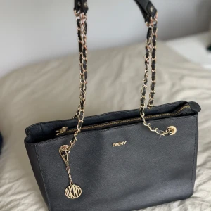 Svart axelväska från DKNY  - Snygg svart axelväska från DKNY i skinn med guldfärgade kedjor och detaljer.
