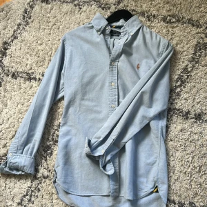 Ljusblå skjorta från Polo Ralph Lauren - Säljer en ljusblå skjorta från Polo Ralph Lauren i slim fit. Skjortan har klassisk krage, knappar framtill och broderad logga på bröstet. Tillverkad i mjuk bomull och har långa ärmar. Perfekt för en clean och stilren look.
