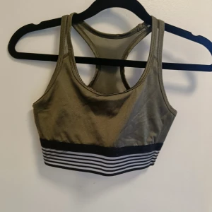 Olivgrön sportig linnetopp - Snygg olivgrön linnetopp med svarta och vita ränder längst ner. Modellen är ärmlös med brottarrygg och passar perfekt till träning eller chill. Materialet är stretchigt och bekvämt, vilket gör den enkel att röra sig i.