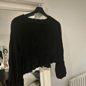 Svart blus med fint mönster och volang - Säljer en svart blus med långa ärmar, knappar framtill, volang vid magen och ryggen och ett fint mönster vid volangen! Buda 🫶🏻🫶🏻
