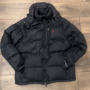  Polo Ralph Lauren - Säljer en svart pufferjacka från Polo Ralph Lauren med röd broderad logga på bröstet. Jackan har hög krage, dragkedja framtill och flera fickor med dragkedja. Perfekt för kalla dagar och riktigt snygg streetstil. Den är äkta köpt från zoovilage 