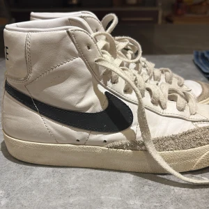 Nike Blazer Mid 77 Vintage Vit/Svart - Nike Blazer Mid 77 Vintage i vitt skinn med svarta swoosh-loggor och beige mockadetaljer runt tån. Klassisk high-top siluett med snörning och retrovibbar. 