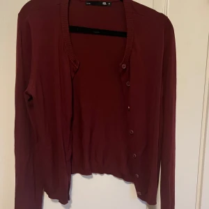 Vinröd kofta från 157, storlek M - Enkel och snygg vinröd kofta från Lager 157 i storlek M. Koftan har långa ärmar, ribbad kant vid halsen och knäppning framtill med matchande knappar. Perfekt att slänga över en t-shirt eller linne. Enbart använd ett par gånger.