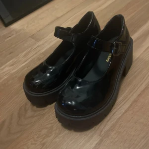 Svarta Mary Jane-plattformskor strl 37 - Chunky Mary Jane-inspirerade skor i svart lack med bred plattformsula och hög klack. Rem med spänne över foten och rund tå. 