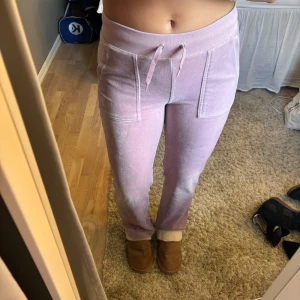 Ljuslila/rosa mjukisbyxor från Juicy Couture - Säljer ett par ljuslila/rosa mjukisbyxor från Juicy Couture med bred resår och snörning i midjan. Byxorna har raka ben, stora fickor fram och en ficka bak med knappar. Mjukt velourmaterial och broderad JC-logga på ena fickan.