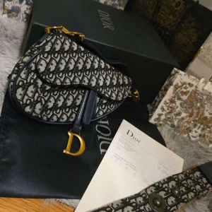 Dior Saddle Bag i blått canvas - Ikonisk Dior Saddle Bag i blått och beige canvas med monogrammönster, detaljer i mörkblått skinn och guldiga metalldetaljer. Väskan har den klassiska D-formade spännet framtill och justerbar axelrem. Perfekt statement piece för dig som älskar lyxiga accessoarer.