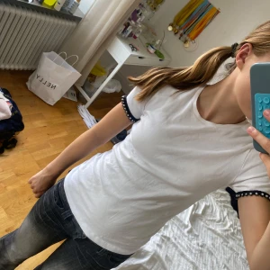 Vit t-shirt med pärldetaljer från Zara - Vit t-shirt från Zara i mjuk bomull med korta ärmar. Ärmsluten har svarta spetsdetaljer och pärlor som ger en unik och snygg touch. Passformen är normal och t-shirten har rund hals. Perfekt för dig som gillar stilrena plagg med coola detaljer.