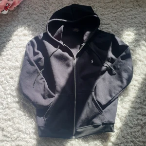 Svart hoodie från Polo Ralph Lauren - Svart hoodie från Polo Ralph Lauren med klassisk broderad logga på bröstet. Tröjan har huva med snörning, dragkedja framtill och två fickor. Materialet är mjukt och känns som bomull, perfekt för chill dagar. Snygg och enkel design som funkar till det mesta. Skriv för mera information  lapp finns kvar med 