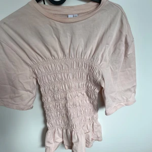 Ljusrosa smockad topp från & Other Stories - Supersöt ljusrosa topp från & Other Stories med smockad midja och volang nertill. Toppen har korta ärmar och är gjord i mjuk bomull. Perfekt för dig som gillar romantiska och feminina detaljer.