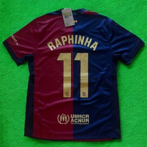 FC Barcelona Raphinha 11 matchtröja Nike - Snygg FC Barcelona matchtröja med Raphinha 11 på ryggen. Tröjan är kortärmad och har en delad design i vinrött och marinblått med guldiga detaljer, klubbmärke och sponsorer. Tillverkad i lätt och ventilerande polyester, perfekt för fotboll eller att bära som supporter.