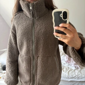 Brun teddyjacka med dragkedja - Mysig brun teddyjacka med hög krage och hel dragkedja framtill. Jackan har två fickor med detaljer i samma färg och en loose passform. Perfekt för kyliga dagar när du vill vara både varm och stilren. Perfekt nu till hösten 