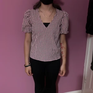 Rutig rosa blus från Bubbleroom - Supersöt rutig blus från Bubbleroom i rosa och svart. Blusen har puffiga korta ärmar, v-ringad hals och en loose passform. Materialet känns lätt och skönt mot huden, perfekt för dig som gillar en chill men ändå trendig look.