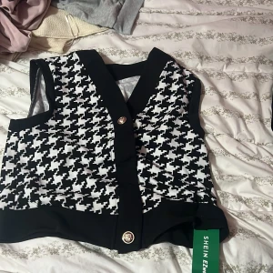 Svartvit rutig väst från SHEIN - Snygg väst från SHEIN i svartvitt rutigt mönster med breda svarta kanter och dekorativa knappar framtill. Västen har en klassisk v-ringning och är tillverkad i ett mjukt syntetmaterial. Perfekt för att lyfta din outfit med en trendig touch.