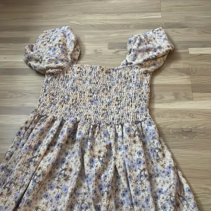 Blommig kortklänning från H&M - Supersöt kortklänning från H&M med puffärmar och smockad överdel. Klänningen har ett blommigt mönster i ljusa toner med inslag av blått och beige. Perfekt för sommarens alla tillfällen, med en luftig och bekväm passform.