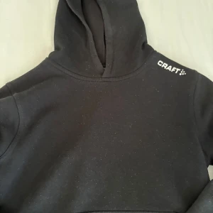 Svart hoodie från Craft, junior - Svart hoodie från Craft med stilren design och logga på axeln. Tröjan har huva och är långärmad, perfekt för chill dagar eller träning. Mjuk insida och klassisk passform. Snygg att matcha med jeans eller joggers.