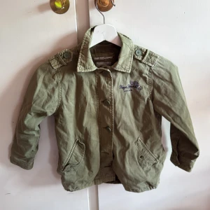 Grön jacka från Pepe Jeans med broderi - Snygg militärgrön jacka från Pepe Jeans med broderade blommor och logga på ryggen. Jackan har knappar framtill, två fickor och coola axeldetaljer. Perfekt för dig som gillar streetstyle och vill sticka ut med blommigt broderi.