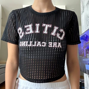 Svart mesh croppad topp med tryck - Säljer en svart croppad mesh-topp med coolt hålmönster och trycket 'CITIES ARE CALLING' i vitt och rött på bröstet. Toppen har korta ärmar och rund hals, perfekt för att ge din outfit en edgy vibe.