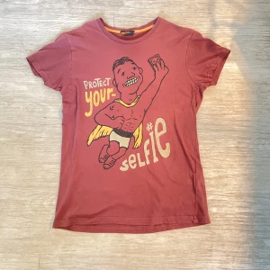 Röd graphic tee från FSBN  - Cool röd t-shirt från FSBN med tryck av en tecknad superhjälte och texten 'Protect your selfie'. T-shirten har rund hals, korta ärmar och är gjord i mjuk bomull. Perfekt för dig som gillar roliga prints och chill stil.