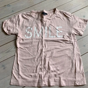 Ljusrosa t-shirt med SMILE-tryck - Söt ljusrosa t-shirt från XOXO Friends med stort vitt SMILE-tryck på bröstet. Klassisk rund halsringning och korta ärmar. Tillverkad i mjuk bomull som är skön att ha på sig hela dagen.
