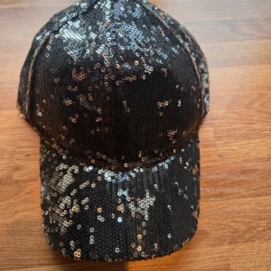 Svart paljettkeps från Lindex - Snygg svart keps från Lindex täckt med glittriga paljetter som ger en riktigt cool och festlig vibe. Justerbar baktill för perfekt passform. Perfekt för dig som vill sticka ut och addera lite extra bling till din outfit.