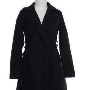 Svart kappa från Amisu, EU 34 - Stilren svart kappa från Amisu i klassisk modell med markerad midja och långa ärmar. Jackan har bred krage, diskreta fickor och snygga detaljer vid sidorna. Perfekt för dig som gillar en clean och tidlös look. Materialet är polyester.