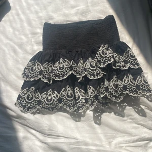 Svart volangkjol med broderi 💞 - Supersöt svart kortkjol med bred smock i midjan och två lager volanger. Kjolen har vita broderade detaljer i ett unikt mönster längs kanterna som ger en extra cool touch. Perfekt för dig som gillar att sticka ut med din stil.