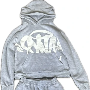 Grå tracksuit ifrån Syna world - Grå mjukisset i storlek S, knappt använt och i superbra skick! Hoodie med magficka och tryck framtill, samt matchande flared byxor med justerbar midja. Perfekt både för mys hemma och en streetwear stil utomhus. Med märket synaworld.