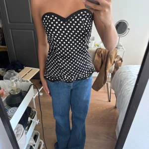 Prickig tubtopp från Pinky Girls - Snygg svart tubtopp med vita prickar från Pinky Girls. Toppen har en söt volangkant nertill och hjärtformad urringning. Den är axelbandslös och har en figurnära passform, perfekt för att sticka ut lite extra. Materialet känns mjukt och glansigt. Passar xs-s.