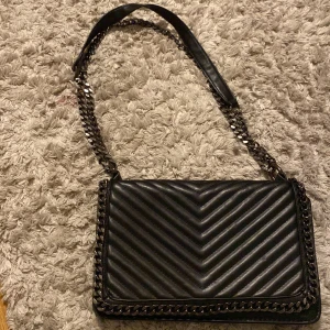 Svart axelväska Gina Tricot - Snygg svart axelväska från Gina Tricot med quiltad design i chevronmönster. Väskan har en kedjerem med läderdetalj upptill och metallkedja runt kanterna för en edgy look. Perfekt storlek för det viktigaste och stängs med lock.