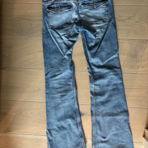 Blå bootcut jeans med dubbla knappar - Snygga blå jeans med bootcut passform och coola slitningar. Byxorna har dubbla knappar i midjan och klassiska fickor både fram och bak med dekorativa detaljer. Perfekta för en avslappnad och trendig look.
