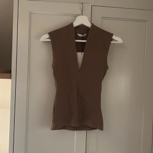 Brun ribbad ärmlös topp från Zara - Säljer en brun ribbad topp från Zara med djup v-ringning och ärmlös design. Toppen har en slim passform och är tillverkad i ett stretchigt material som sitter snyggt på kroppen. Perfekt att styla med jeans eller kjol för en trendig look. Lappen kvar!