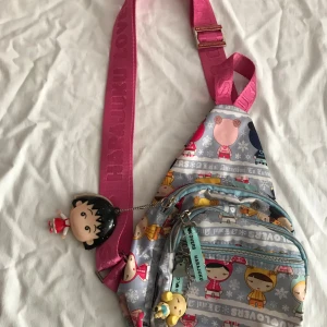 Harajuku Lovers ryggsäck med figurmönster - Superfin ryggsäck från Harajuku Lovers med färgglada figurer på grå bakgrund och snöflingor. Axelremmen är bred och rosa med logga, och väskan har flera dragkedjor samt söta nyckelringar. Perfekt för dig som gillar lekfull stil och japansk popkultur. En dragkedja är trasig med spegel 