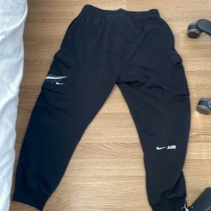 Svarta cargobyxor från Nike Air M - Svarta cargobyxor från Nike Air med elastisk midja och muddar vid benslut. Byxorna har stora fickor på sidorna, Nike Air-logga på benet och är tillverkade i mjuk bomullsblandning. Perfekta för en chill och sportig look.