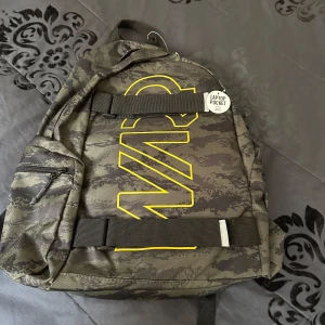 Camouflage ryggsäck med laptopfack - Säljer en snygg ryggsäck från WARP i camouflage-mönster med stora gula bokstäver på framsidan. Väskan har ett separat laptopfack, justerbara axelremmar och en praktisk ytterficka med dragkedja. Perfekt för skolan eller när du är på språng.