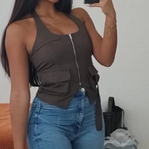 Brun ärmlös linnetopp med fickor - Snygg brun linnetopp med halterneck och dragkedja framtill. Toppen har två stora fickor på sidorna och ett band som knyts i midjan för extra cool stil. Perfekt att matcha med jeans för en trendig look.
