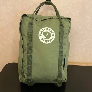 Grön Fjällräven Kånken Tree ryggsäck - Säljer en grön Fjällräven Kånken Tree ryggsäck med klassisk fyrkantig form och stort vitt logotryck framtill. Tillverkad av växtbaserat material, har dubbla handtag upptill, justerbara axelremmar och sidofickor. Perfekt för skola eller cityhängb.• Höjd 38 • Bredd 27 • Djup 14