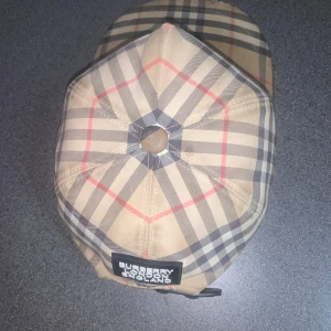 Burberry rutig keps i beige och svart - Snygg keps från Burberry med klassiskt rutigt mönster i beige, svart, vitt och röda linjer. Kepsen har böjd skärm och justerbar rem bak. Märkespatch med Burberry London England baktill. Perfekt accessoar för en trendig look.