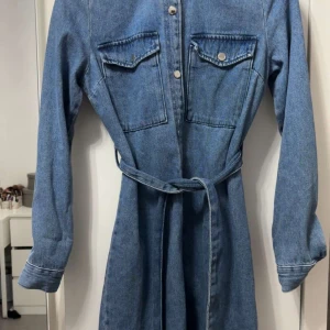 Blå jeansklänning från New Yorker  - Snygg blå jeansklänning från Amisu med långa ärmar, knappar framtill och två bröstfickor med lock. Klänningen har ett matchande bälte i midjan för en cool siluett. Perfekt för dig som gillar denim och vill ha en avslappnad men trendig look.