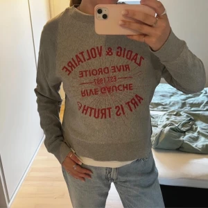 Grå sweatshirt Zadig & Voltaire - Grå sweatshirt från Zadig & Voltaire med röd textprint framtill. Klassisk rund halsringning och långa ärmar. Perfekt till jeans för en avslappnad och cool look.