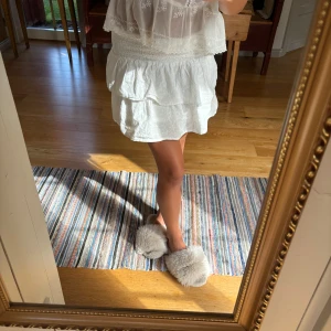 Vit kort kjol med broderade blommor - Supersöt vit kort kjol med volanger och broderade små blommor. Kjolen har en smockad midja som gör den bekväm och sitter snyggt. Perfekt för varma dagar och ger en fräsch och romantisk vibe. Materialet är mjukt och luftigt, troligen bomull.