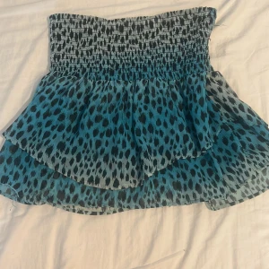 Blå leopardmönstrad kjol från H&M - Supersnygg blå kortkjol från H&M med leopardmönster och volang. Kjolen har en smockad midja som sitter bekvämt och ger en cool siluett. Materialet är lätt och luftigt, perfekt för varma dagar. Passar dig som gillar att sticka ut med djurmönster!